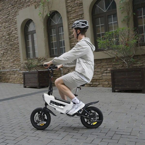Personne faisant du vélo électrique pliable blanc et noir. Porte un casque. Arrière-plan : rue, bâtiments.