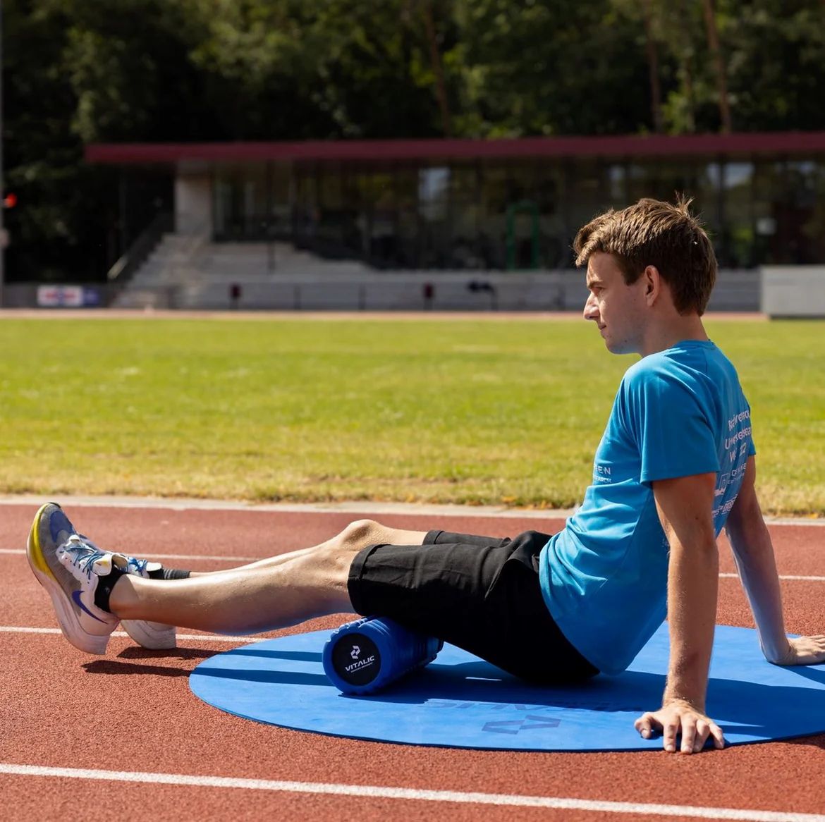 Homme assis sur piste rouge, utilise un rouleau en mousse bleu. Sur tapis bleu.