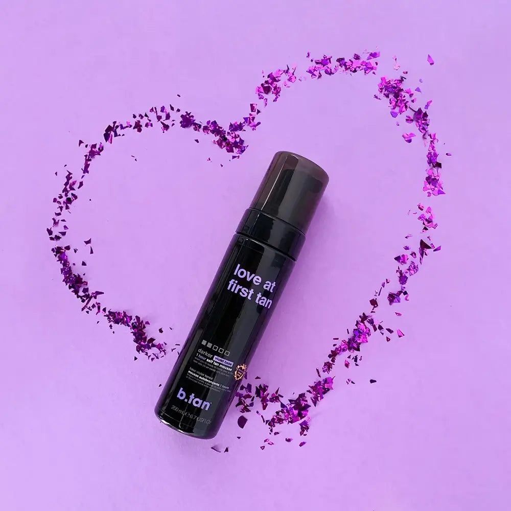 Flacon noir de mousse autobronzante entouré de confettis violets en forme de cœur. Texte: Love at first tan, b.tan.