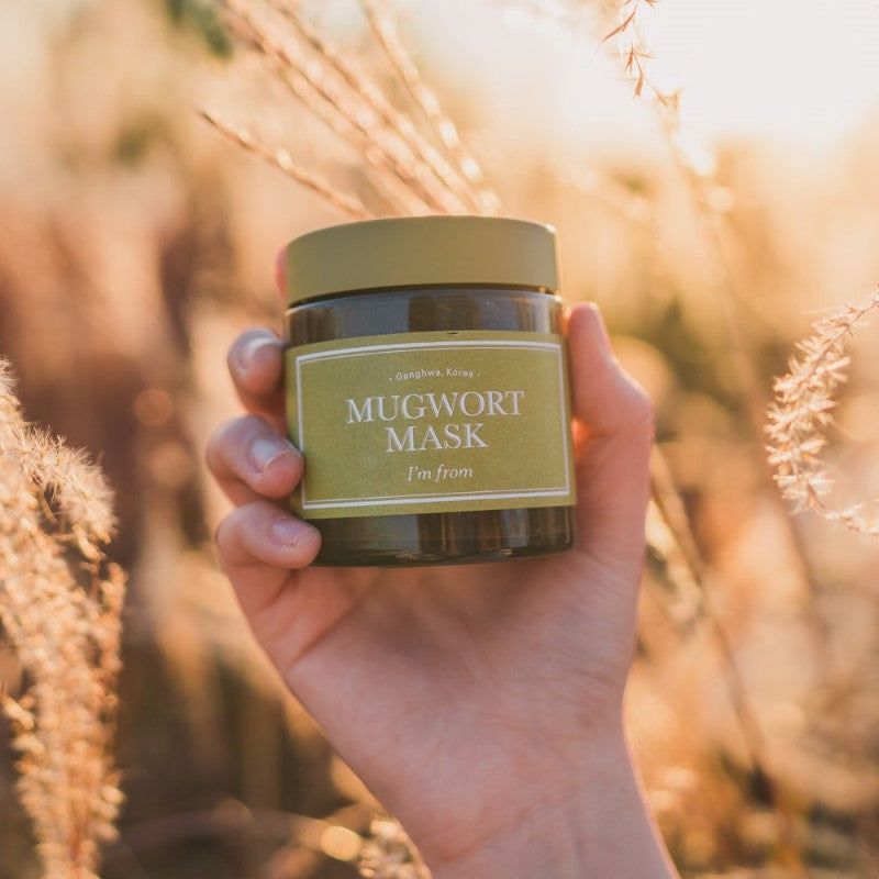 Hand houdt een glazen pot met "Mugwort Mask" etiket. Achtergrond: grassen.