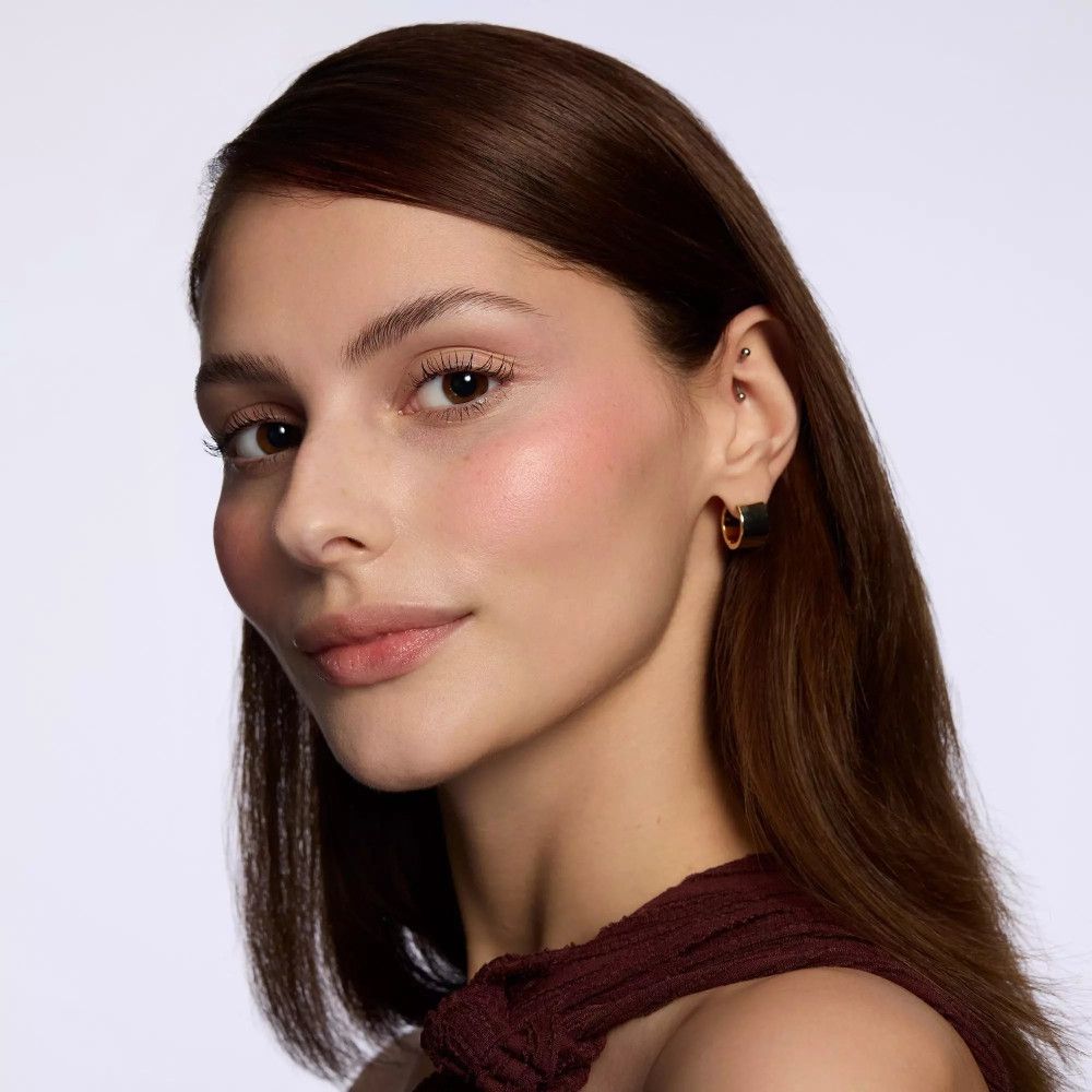 Portrait d'une femme avec du blush rose sur les joues. Elle porte des boucles d'oreilles dorées et a les cheveux bruns.