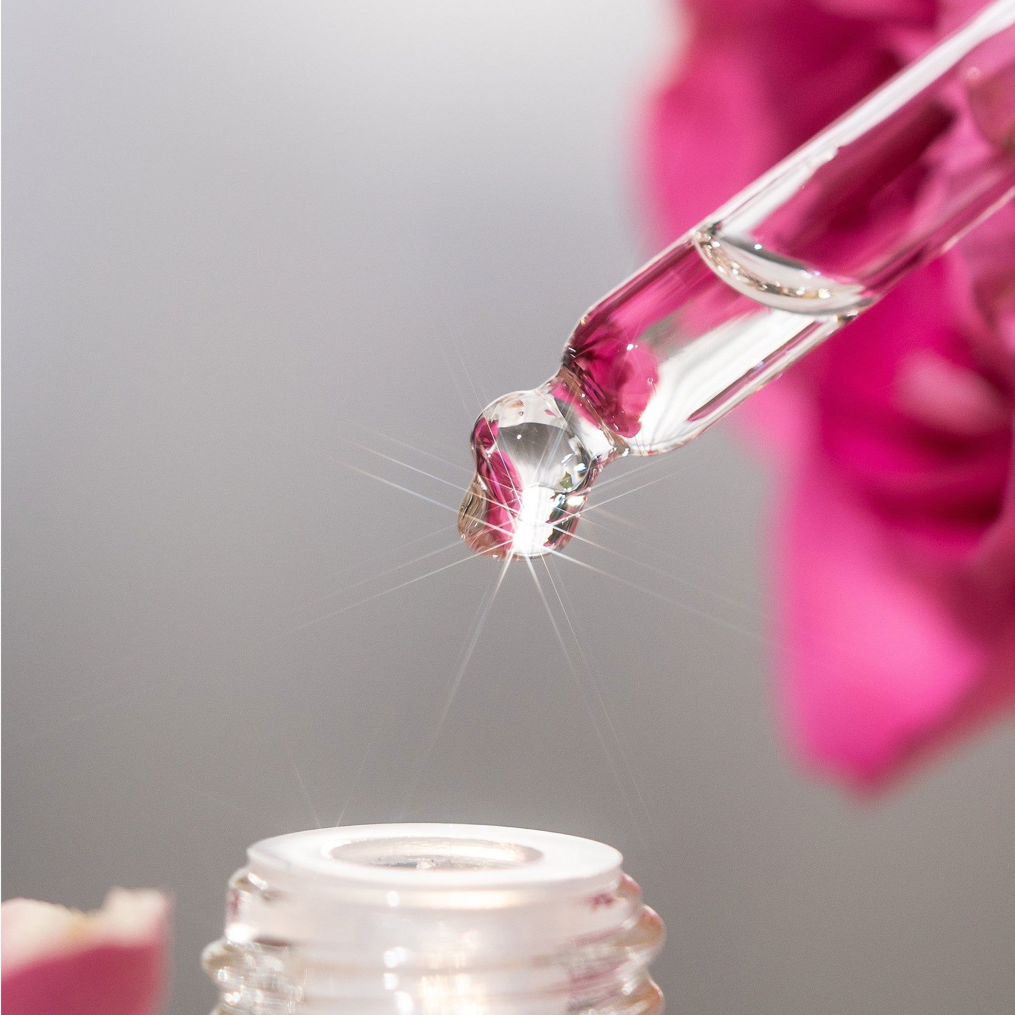 Close-up van een pipet met een druppel. Vloeistof druppelt in een fles. Achtergrond: roze bloem.