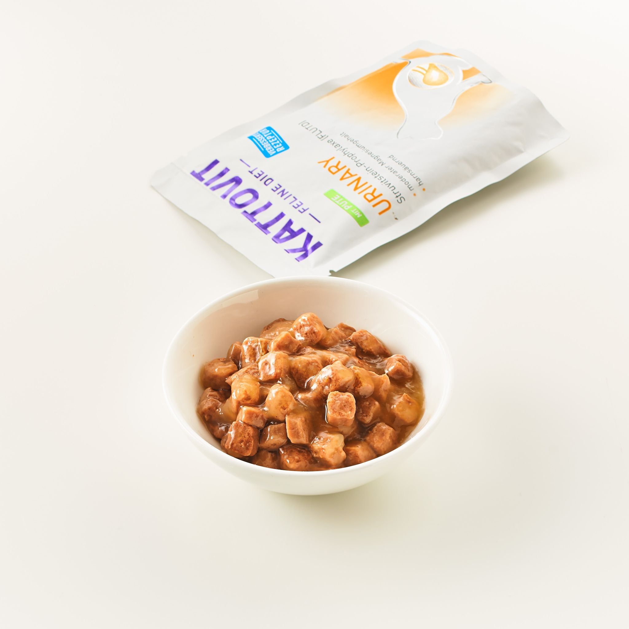 Kom met voedselblokjes naast een KATTOVIT Urinary zakje. Opschrift: KATTOVIT Feline Diet Urinary. Witte achtergrond.