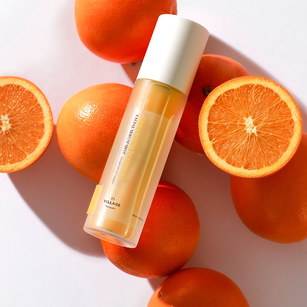 Fles omringd door sinaasappels. Tekst: FACIAL SERUM MIST, -11- VILLAGE FACTORY. Witte dop. Sinaasappels in plakjes.