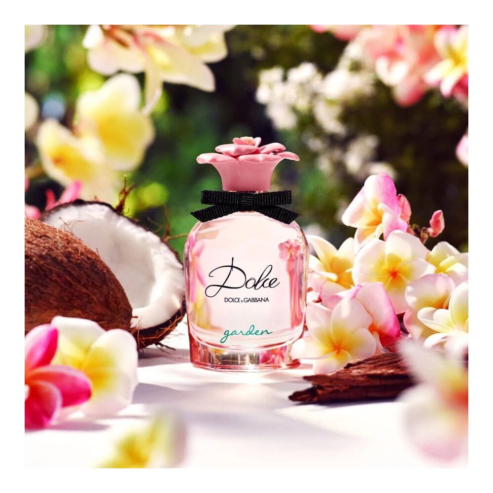Roze parfumfles met bloemvormige dop en zwarte strik. Opschrift "Dolce" en "Dolce & Gabbana". Omringd door kokosnoot en bloemen.