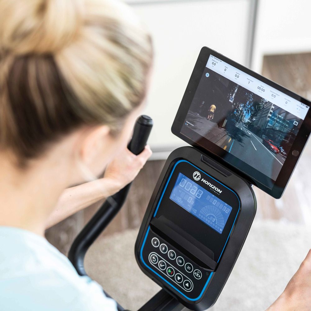 Vrouw traint op een fitnessfiets. Tablet op de console. Horizon Fitness logo. Blauw display met trainingsgegevens.