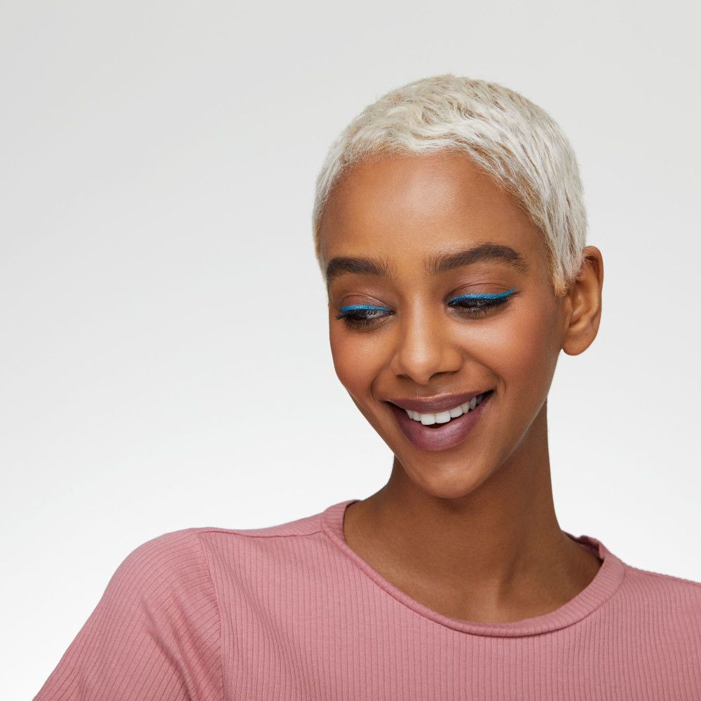 Femme souriante avec eye-liner bleu. Elle porte un haut rose et a les cheveux blonds courts.