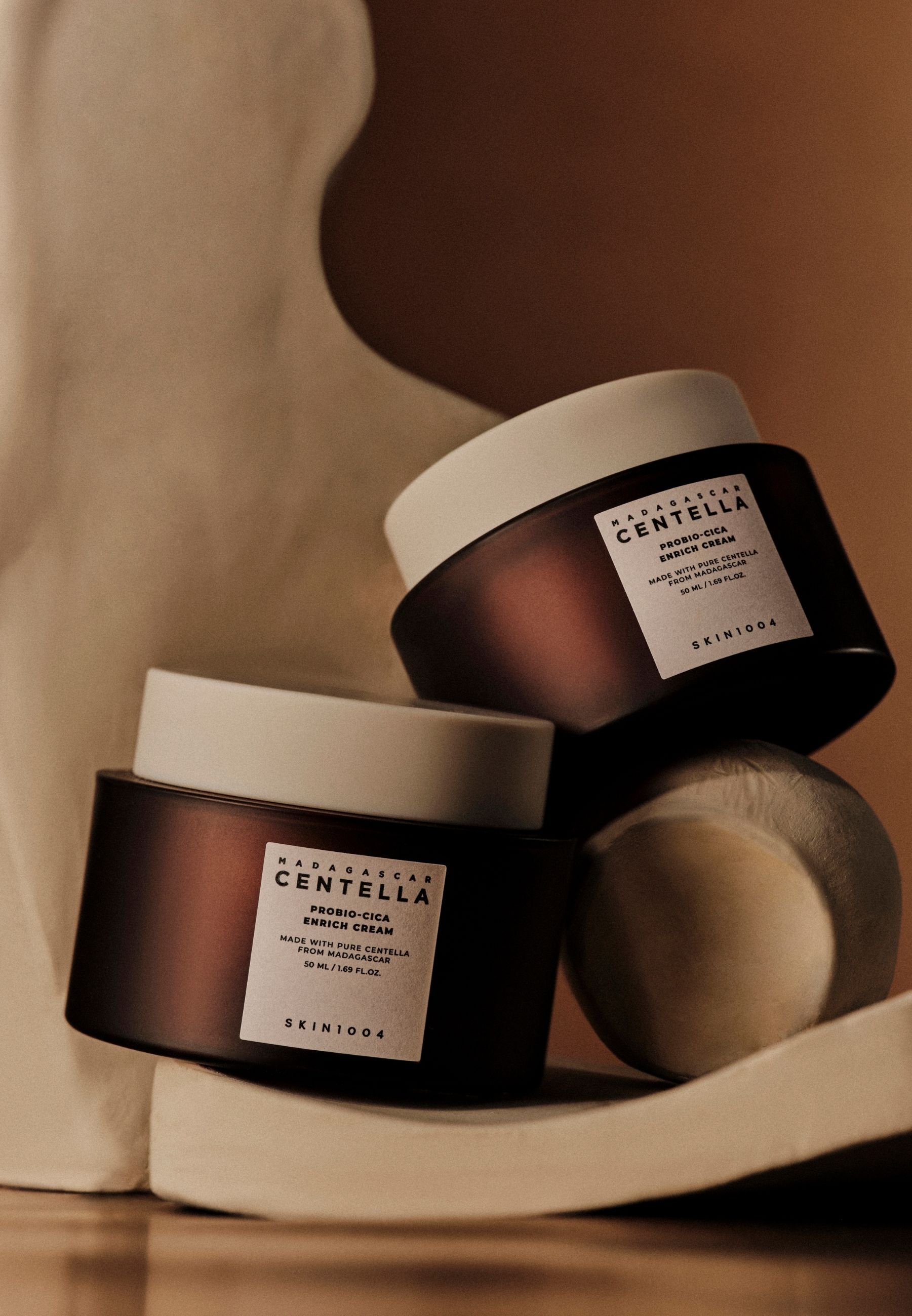 Deux pots de crème bruns avec couvercles blancs, empilés sur des formes abstraites.