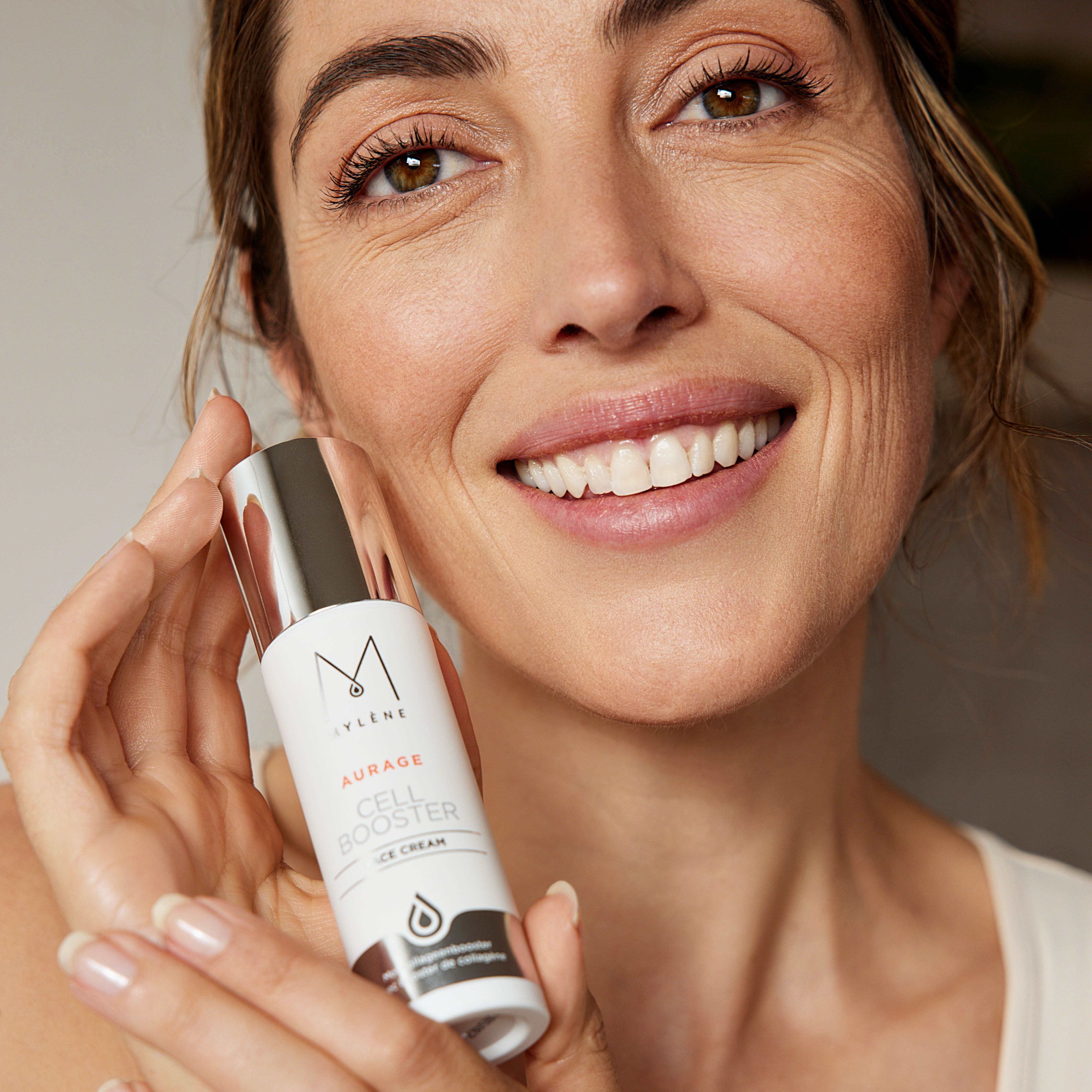 Femme tenant un flacon de crème. Logo MYLÈNE et Aurage Cell Booster. Emballage argenté et blanc. Femme souriante.
