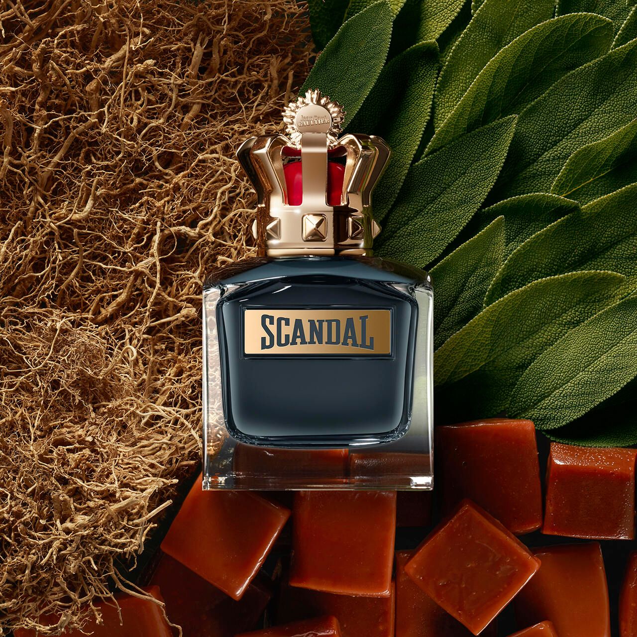 Flacon de parfum avec couronne dorée, entouré d'herbes et de bonbons. Inscription "SCANDAL".