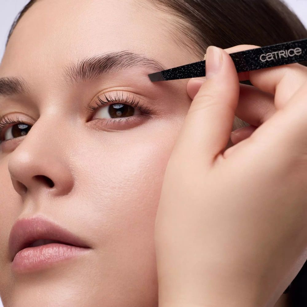 Close-up van een persoon die wenkbrauwen epileert met een pincet. Opschrift: Catrice.