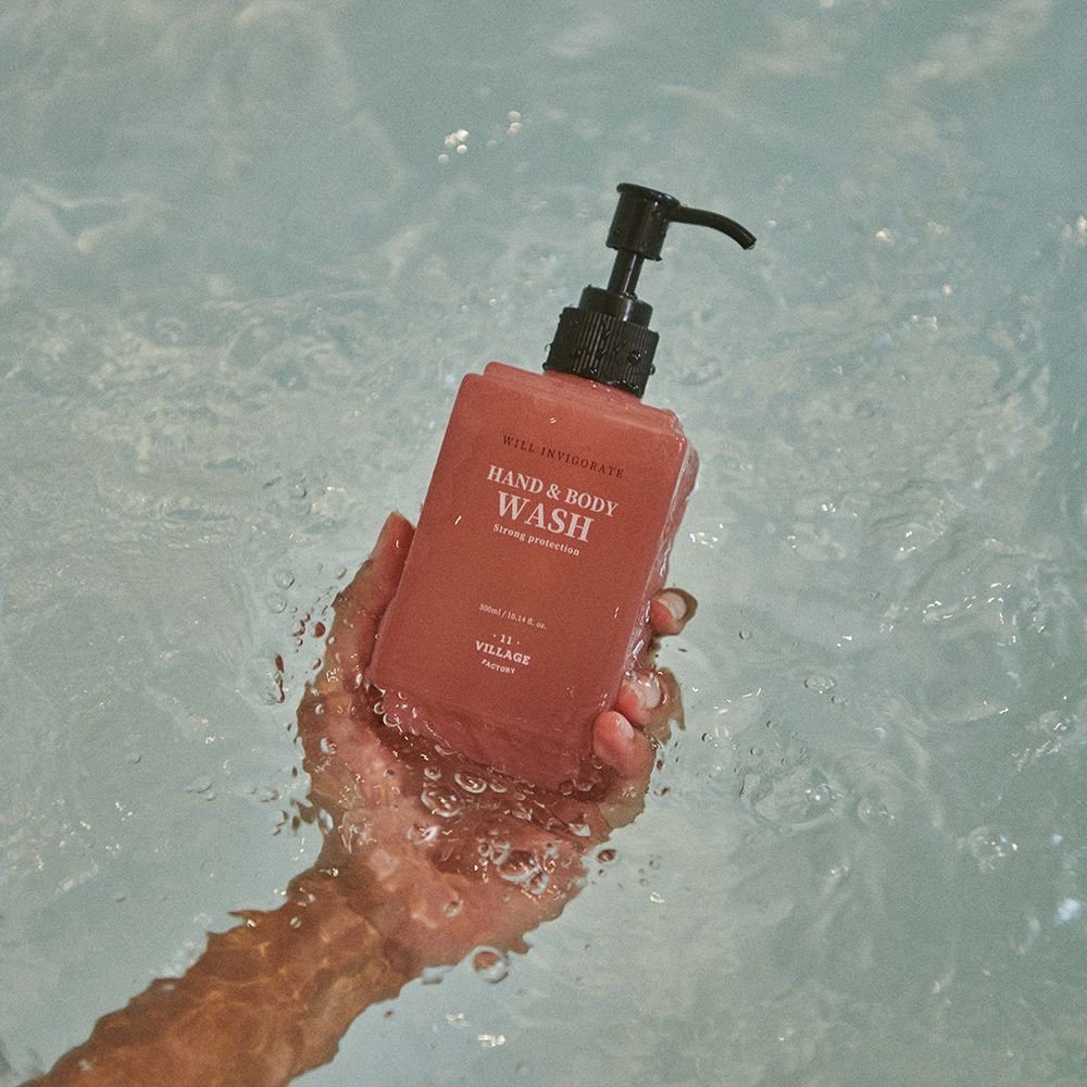 Hand houdt roze hand- en bodywash fles in water. Zwarte pomp. Tekst: HAND & BODY WASH, Strong protection, 11 VILLAGE FACTORY.