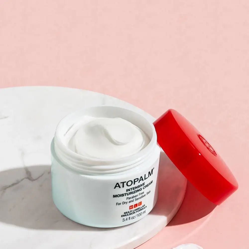 Open crème pot met rode deksel. Opschrift: ATOPALM INTENSIVE MOISTURIZING CREAM. Crème op wit oppervlak.