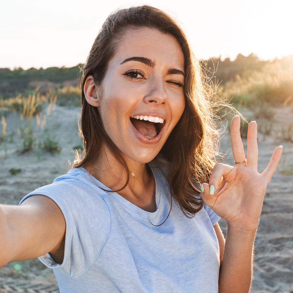 Een vrouw in een blauw T-shirt maakt een selfie. Ze knipoogt en toont het oké-teken.