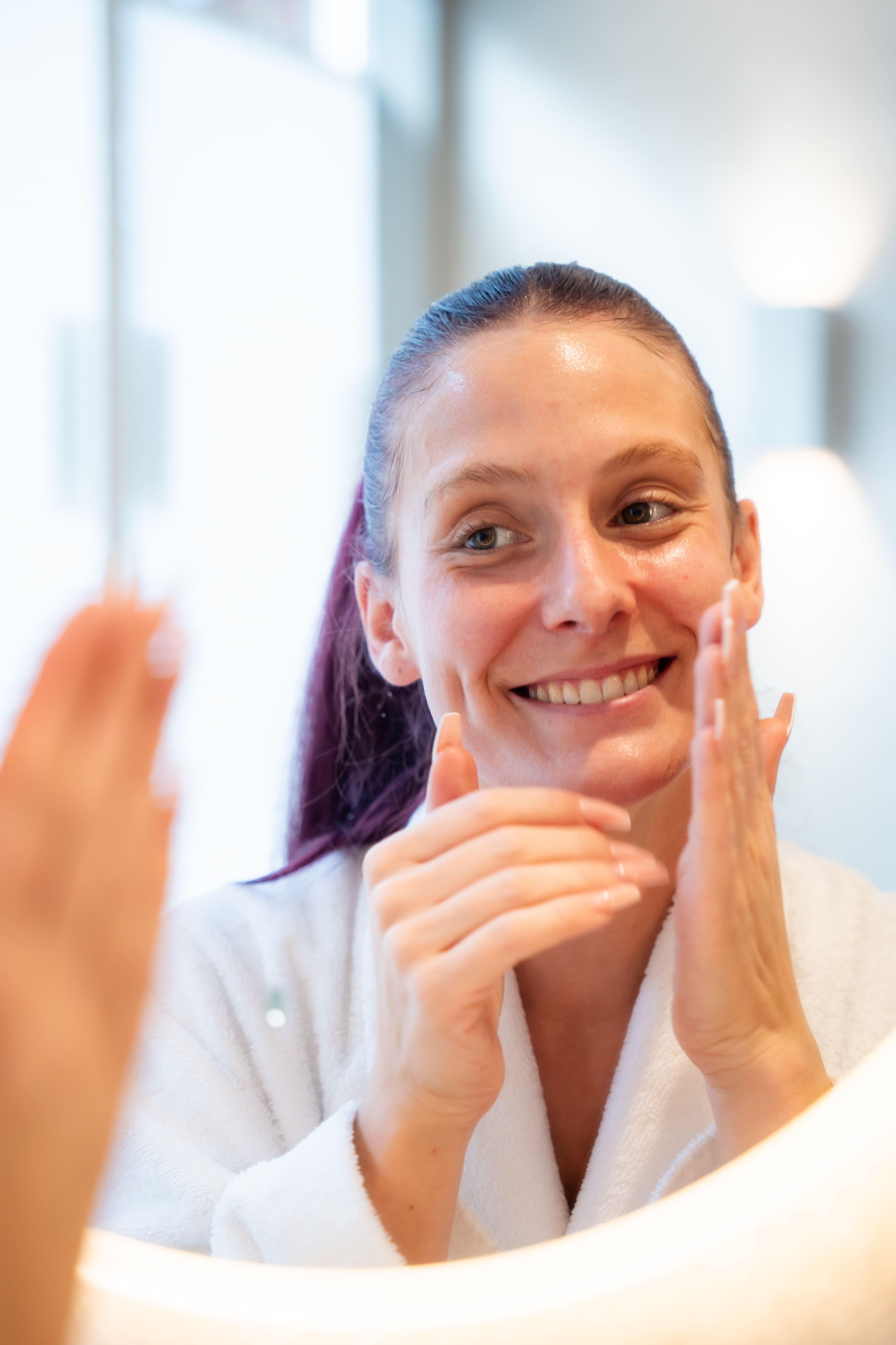 Een vrouw brengt huidverzorgingscrème aan op haar gezicht terwijl ze in een spiegel kijkt.