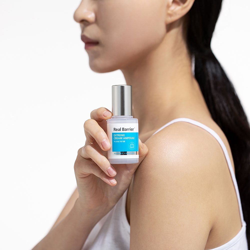 Vrouw houdt flacon vast. Opschrift: REAL BARRIER Extreme Cream Ampoule. Zilveren dop, wit etiket met blauw vlak.