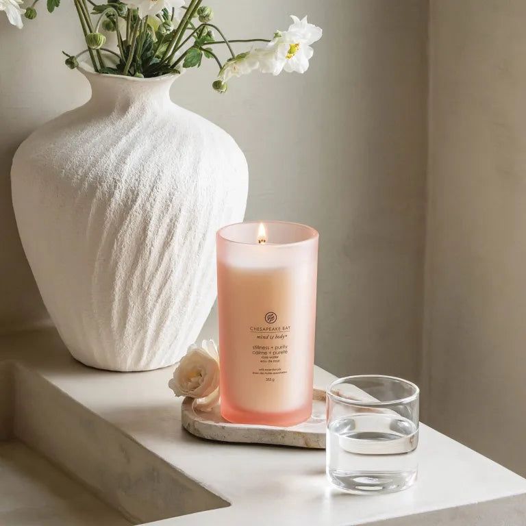 Bougie rose allumée. Inscriptions: Chesapeake Bay, mind & body, stillness + purity, calme + pureté, eau de rose, 355g. Près d'un verre d'eau.