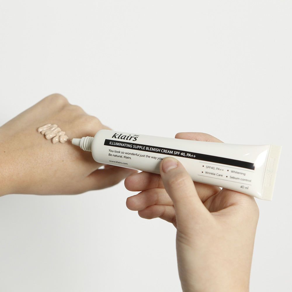 Mains tenant un tube. Crème appliquée sur la peau. Inscription: Dear Klairs Illuminating Supple Blemish Cream SPF 40.