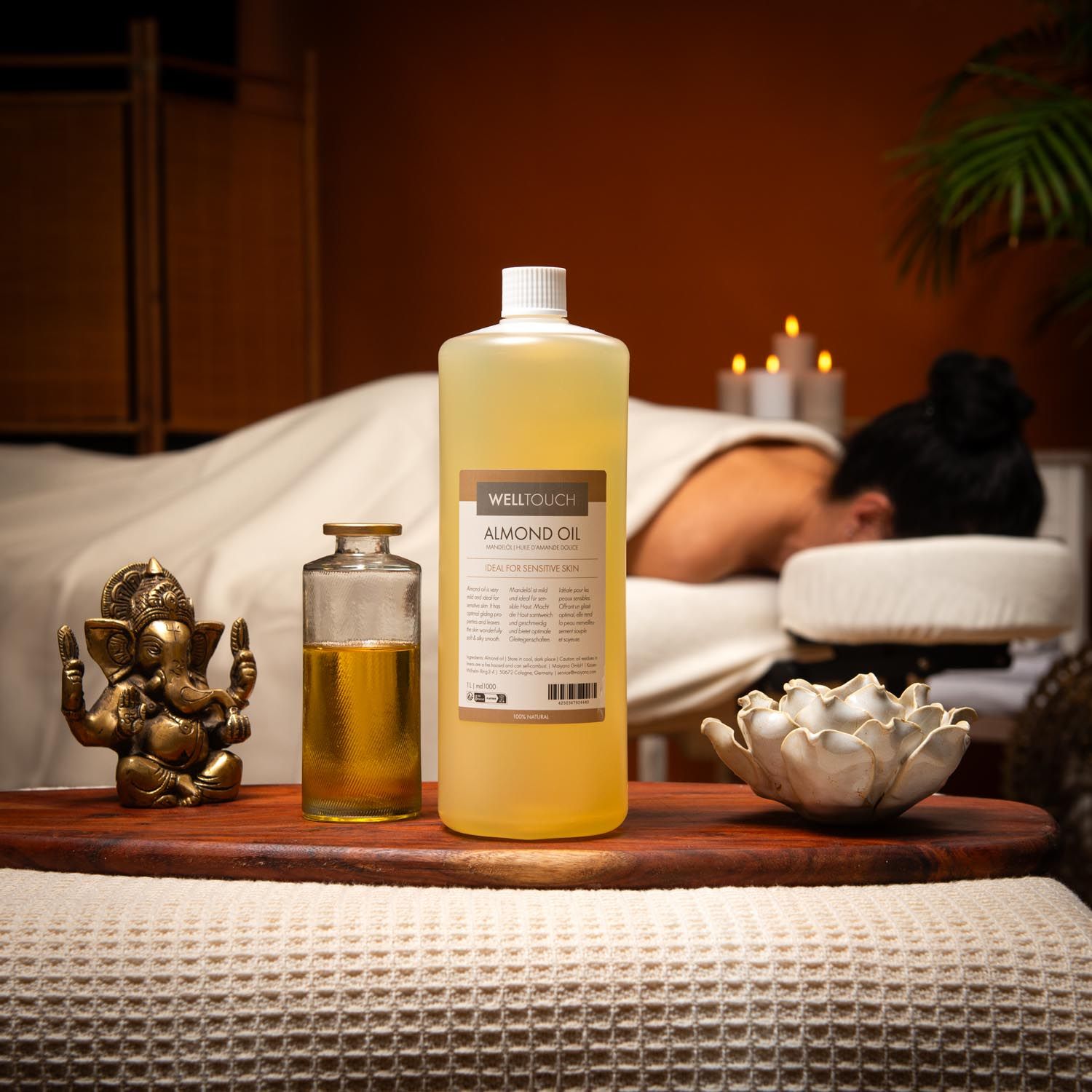 Grote fles en kleine glazen fles met gele vloeistof. Opschrift: Welltouch Almond Oil. Op de achtergrond een persoon op een massagetafel.