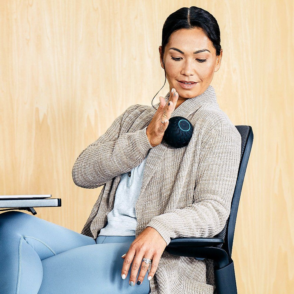 Femme assise tenant la balle de massage noire sur l'épaule. Elle porte un cardigan et un pantalon bleu. Fond en bois.
