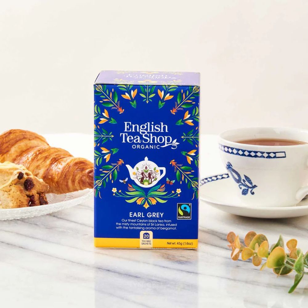 Blauwe doos met opschrift: English Tea Shop, Earl Grey, Organic. Fairtrade-label. Daarnaast gebak en een kopje thee.