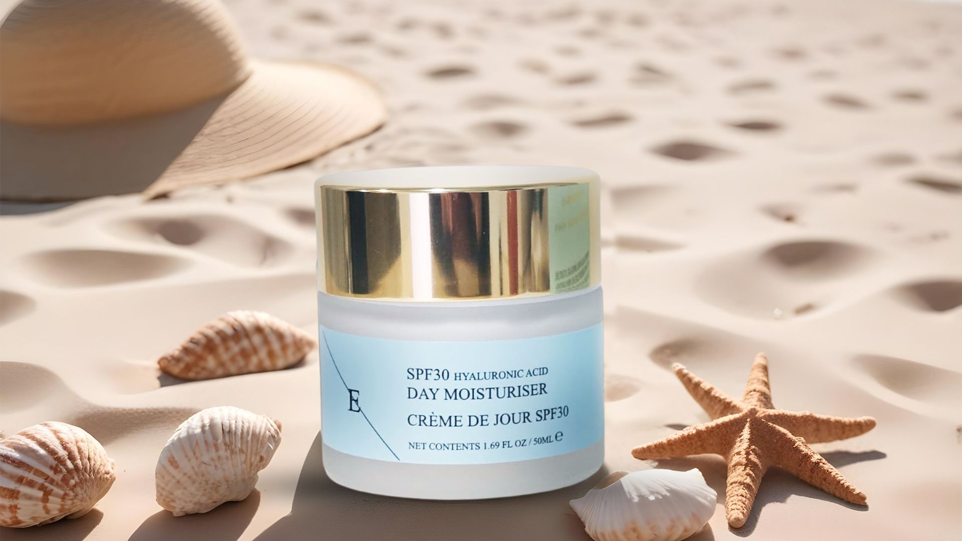 Un pot de crème avec un couvercle doré et une étiquette bleue. Sur le sable, avec des coquillages, une étoile de mer et un chapeau en arrière-plan.