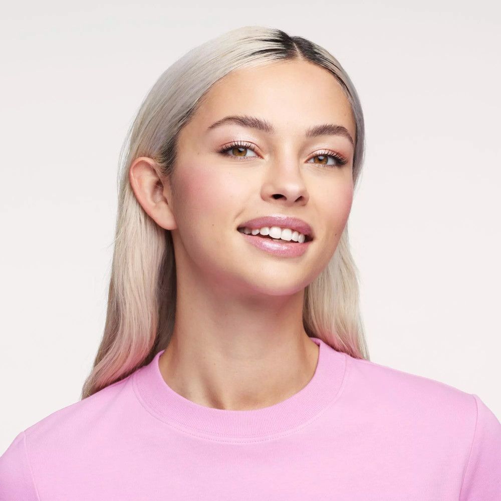 Een vrouw met blond haar en roze lippen. Ze draagt een roze T-shirt en lacht in de camera.