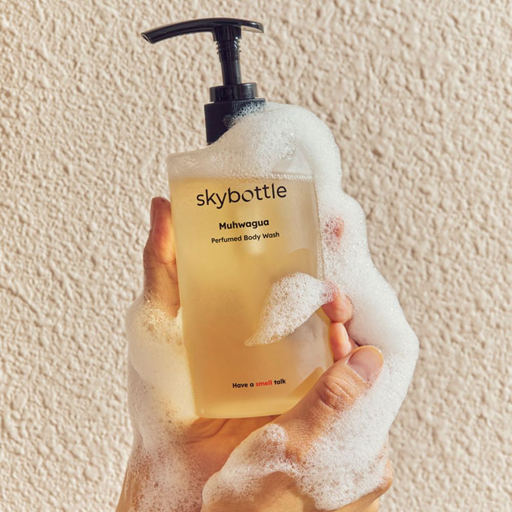 Handen houden een fles met schuim. Opschrift: skybottle, Muhwagua, Perfumed Body Wash. Gele vloeistof.