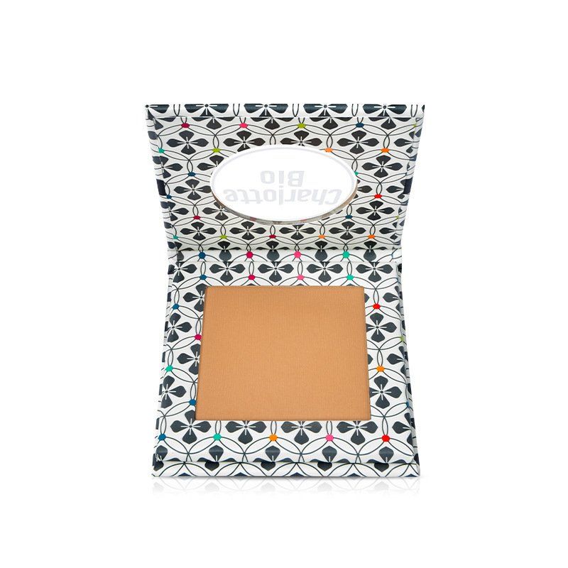 Poudre compacte dans un emballage ouvert. L'emballage est blanc avec un motif floral noir et coloré. La poudre est de couleur dorée.