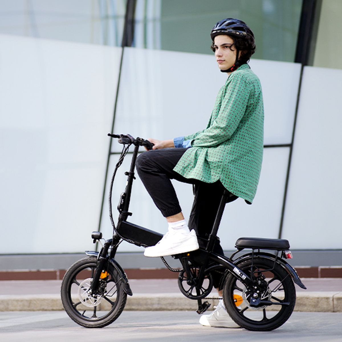 Persoon zit op een zwarte, opvouwbare elektrische fiets. Draagt helm en groene jas. Achtergrond: gebouw.