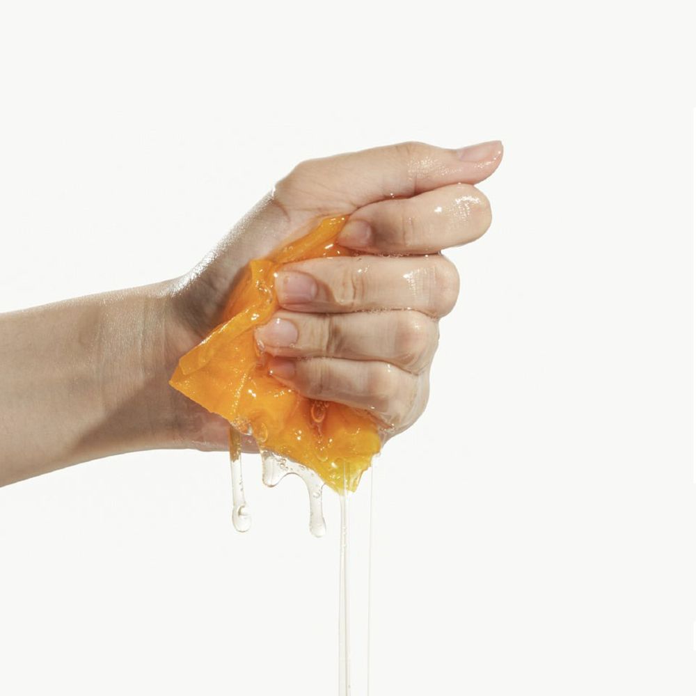 Hand die een geel, doorweekt maskerdoek uitwringt. Vloeistof druppelt. Witte achtergrond.