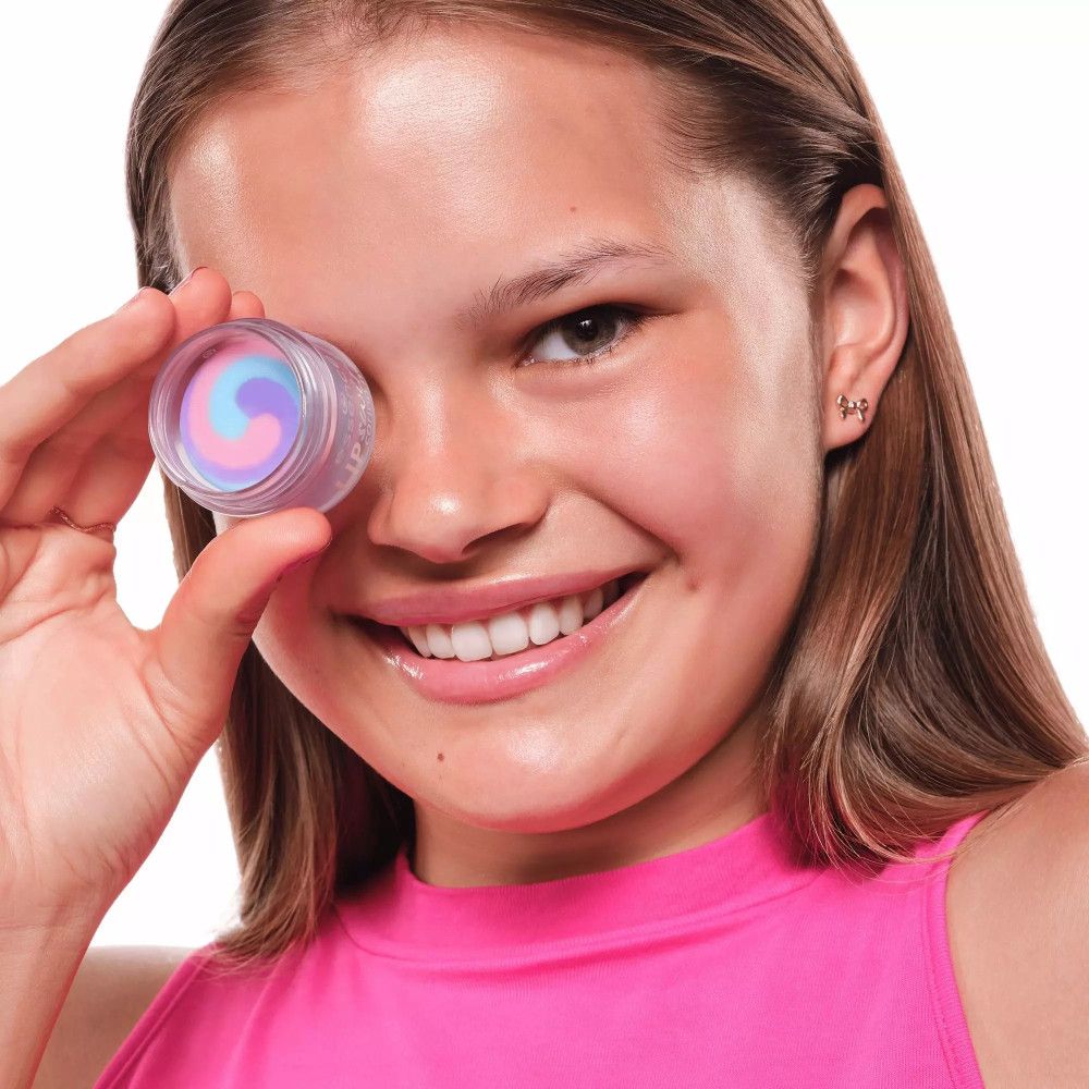 Fille tenant un pot devant un œil. Pot avec des tourbillons colorés. Inscription: essence LIP swirl caring mask.