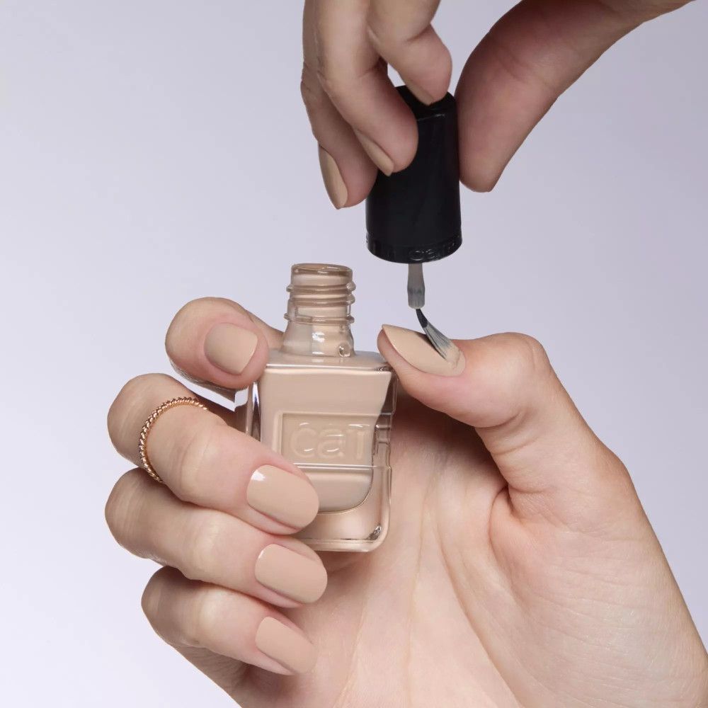 Main avec ongles vernis. Vernis à ongles beige. Flacon de vernis à ongles avec bouchon noir. Logo Catrice.