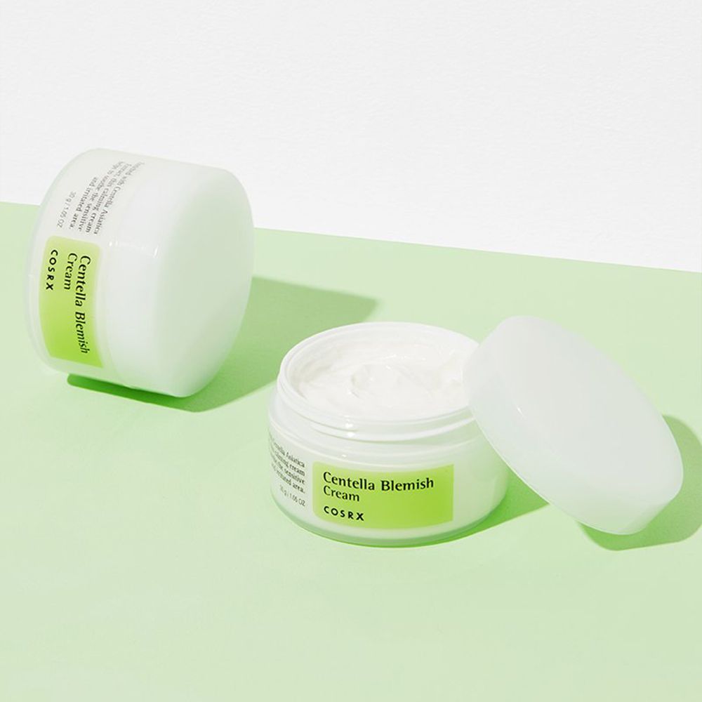 Twee potten COSRX Centella Blemish Cream, één open, op groene ondergrond.