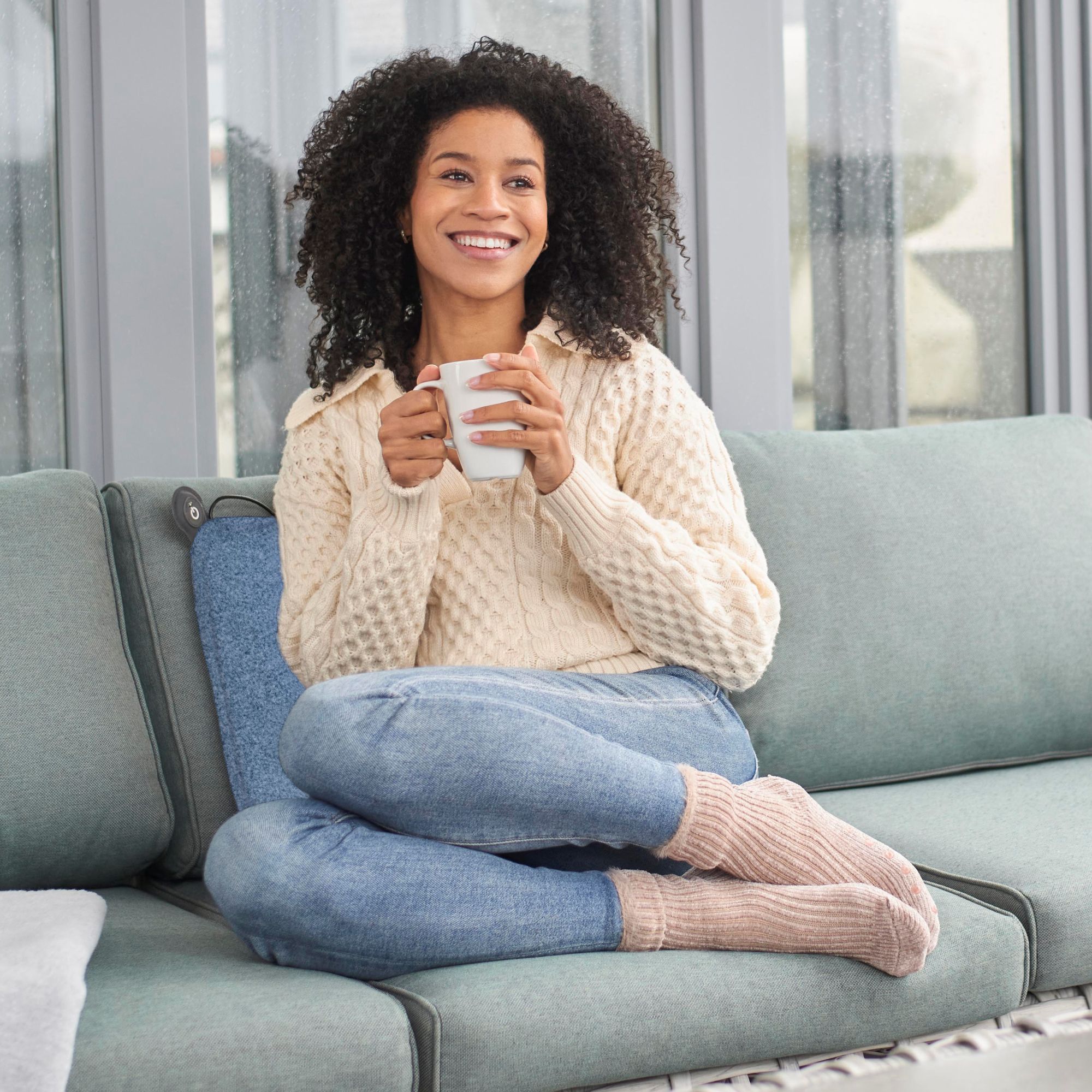 Une femme assise détendue sur un canapé, tenant une tasse, porte un pull et un jean.