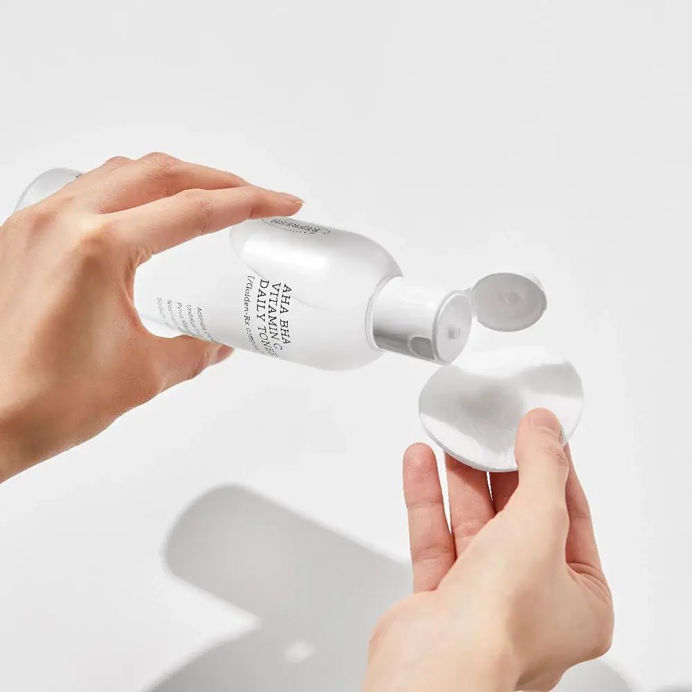 Flacon blanc de toner versé sur un coton. Inscription : AHA BHA Vitamin C Daily Toner.
