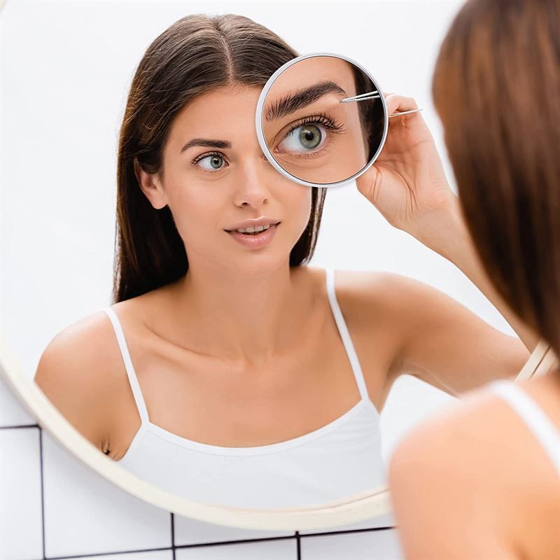 Femme utilisant une pince à épiler pour examiner son œil à travers un miroir grossissant 10x.