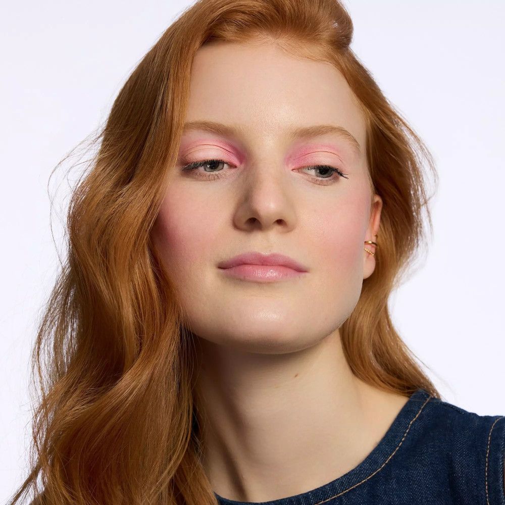 Portrait d'une femme avec fard à paupières rose sur les paupières. Elle a les cheveux roux et porte une chemise en jean.