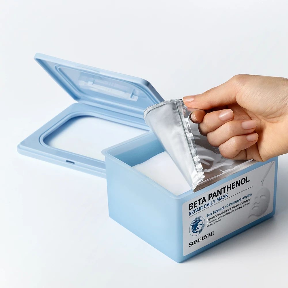 Boîte bleu clair avec couvercle, remplie de masques. Une main ouvre un sachet de masque. Sur la boîte, on lit "Beta Panthenol Repair Daily Mask".