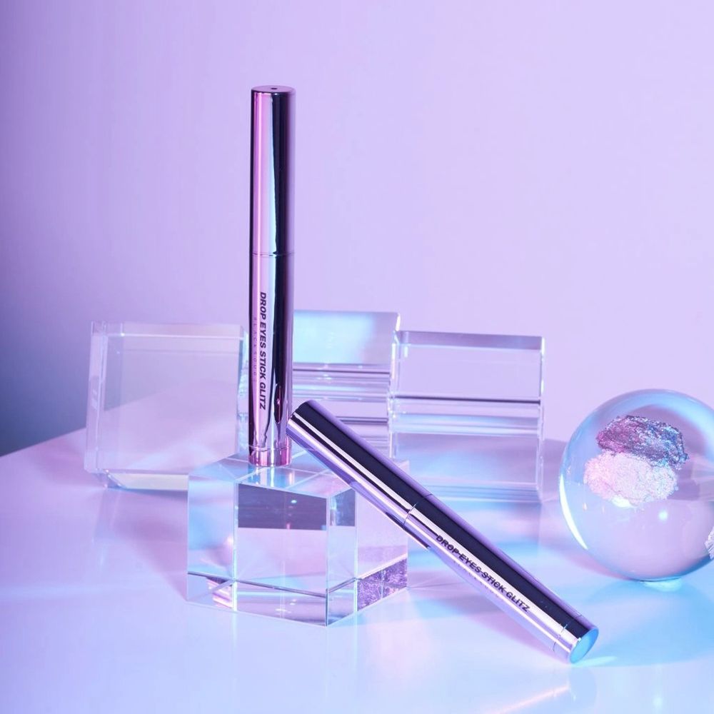 Twee zilverkleurige oogschaduwstiften op sokkels. Opschrift: DROP EYES STICK GLITZ.