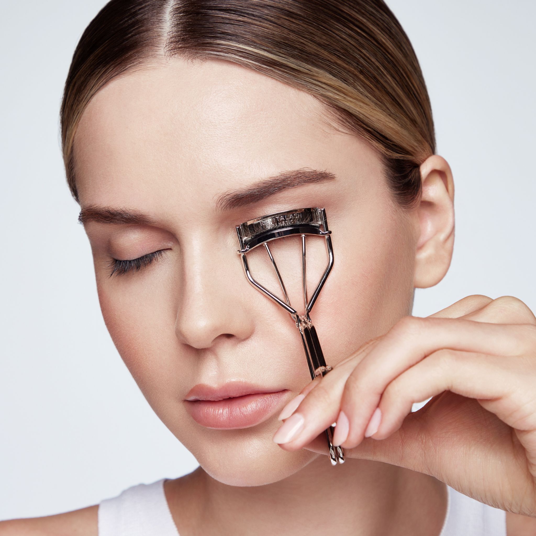 Une femme utilise un recourbe-cils argenté avec un coussinet noir sur son œil gauche.