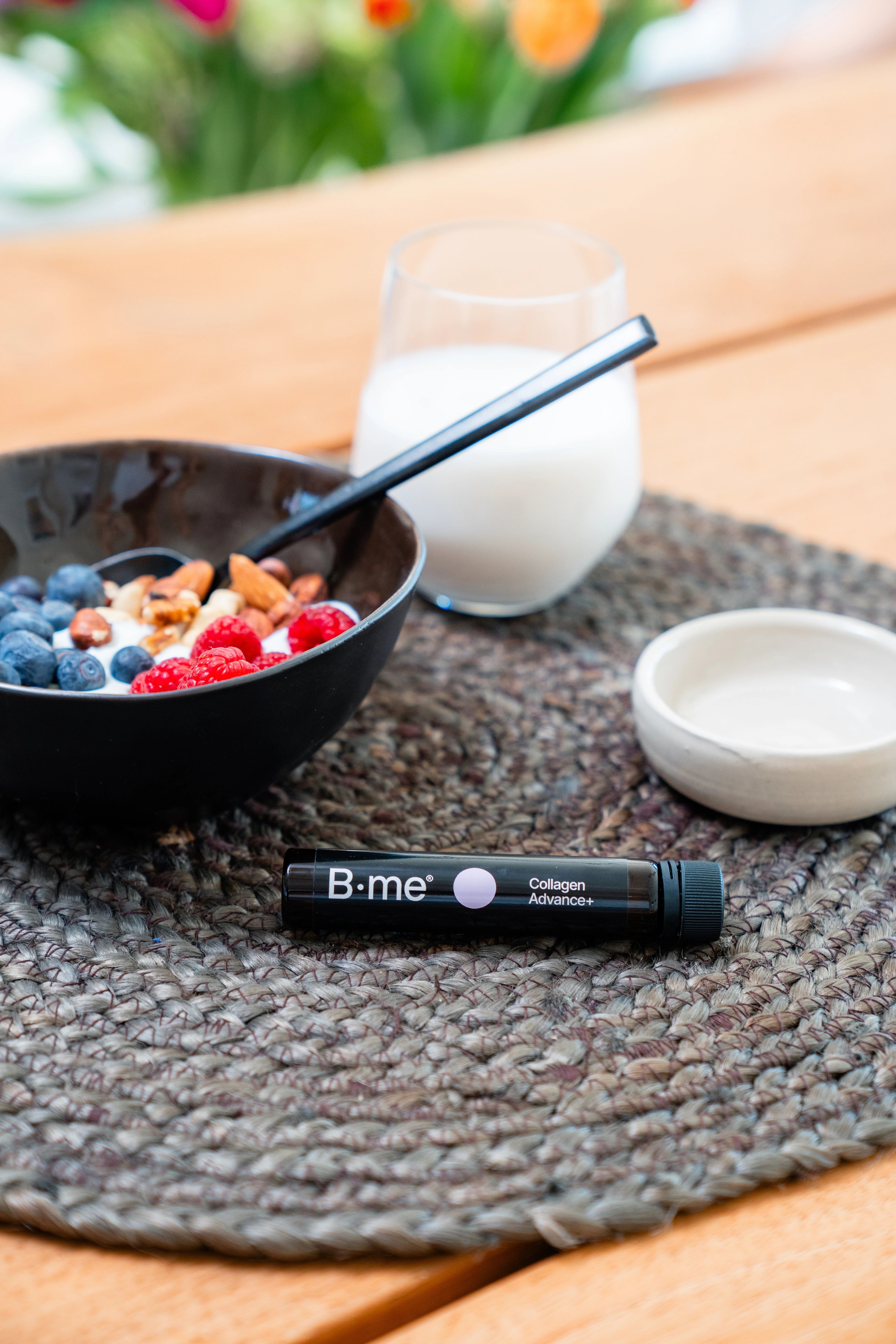 Ampoule B.me® Collagen Advance+ sur une table, à côté d'un bol de nourriture et d'un verre de lait. Bouchon noir, point rose.