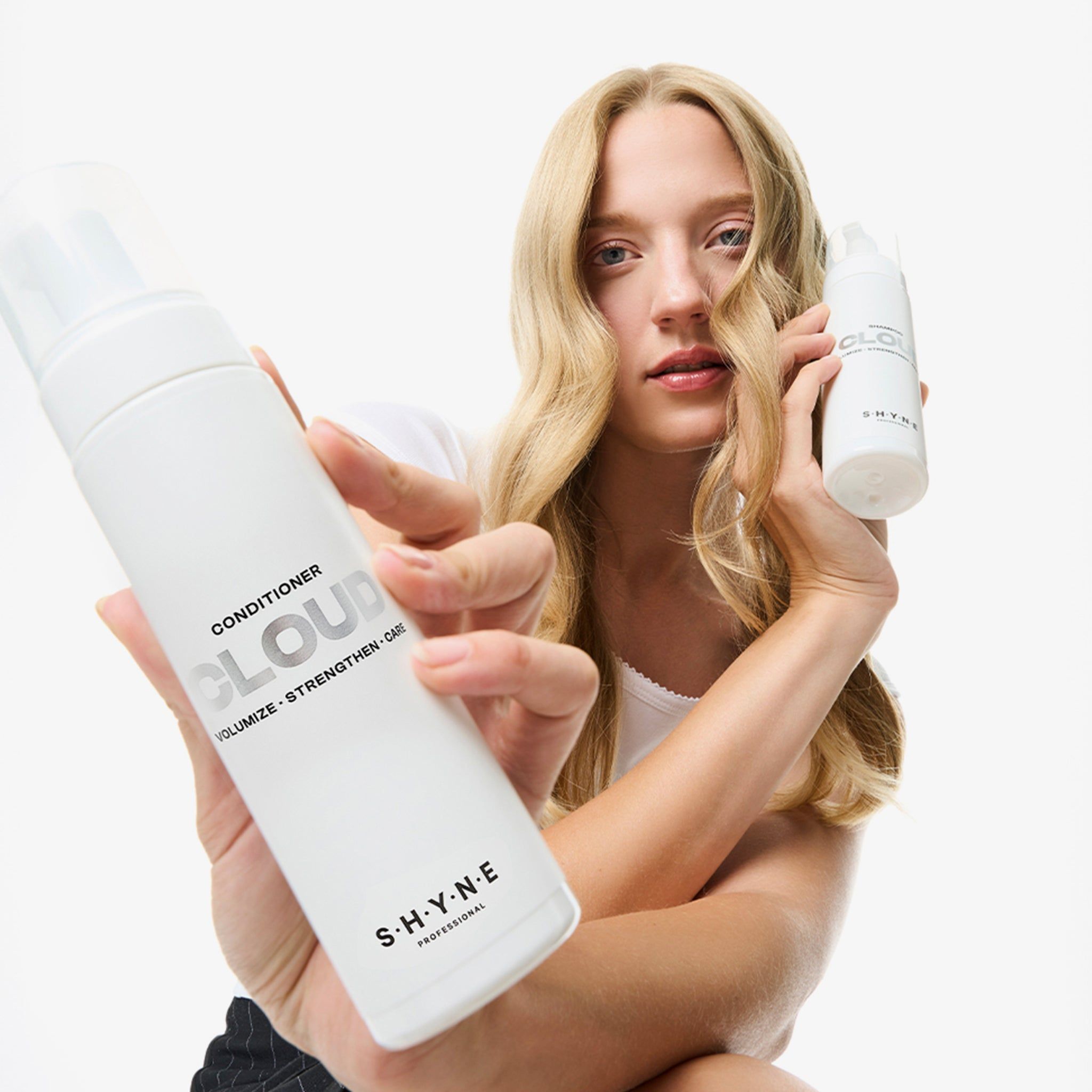 Femme tenant deux flacons blancs. L'un porte l'inscription "CONDITIONER CLOUD" et "S.H.Y.N.E".
