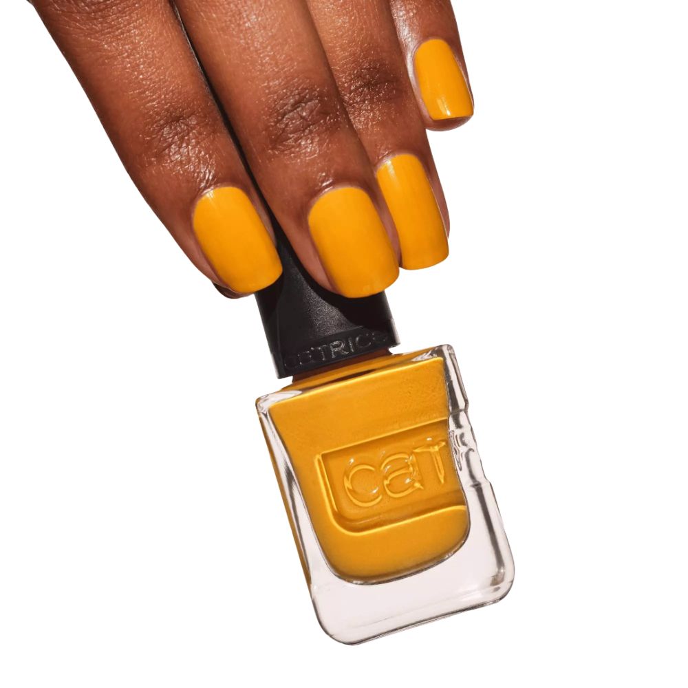 Main avec ongles vernis tenant un flacon de vernis à ongles. Bouchon noir avec inscription Catrice. Vernis à ongles jaune.