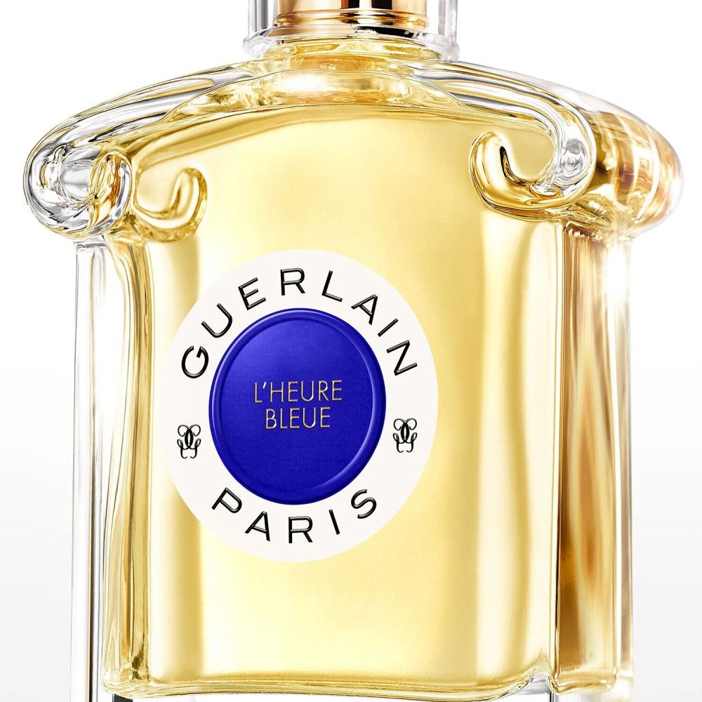 Glazen fles met gele vloeistof. Blauw rond etiket met witte tekst: GUERLAIN PARIS, L'HEURE BLEUE. Twee gouden versieringen.