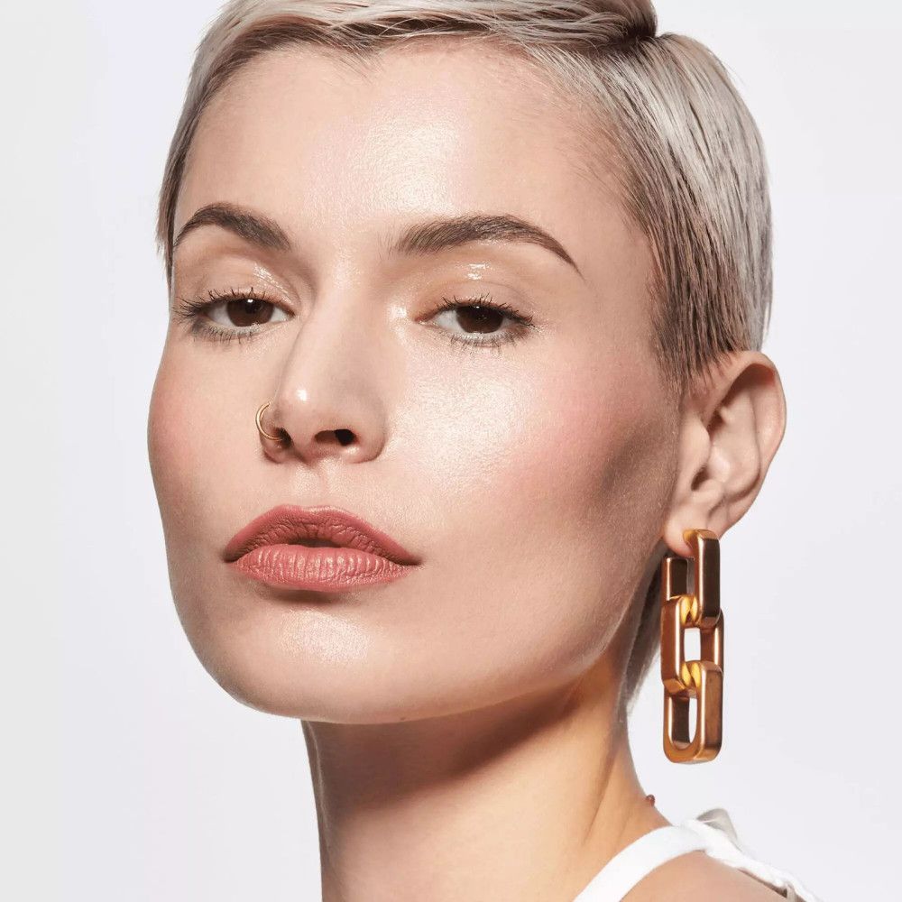 Portrait d'une femme aux cheveux blonds courts. Elle porte du rouge à lèvres rose et des boucles d'oreilles dorées.