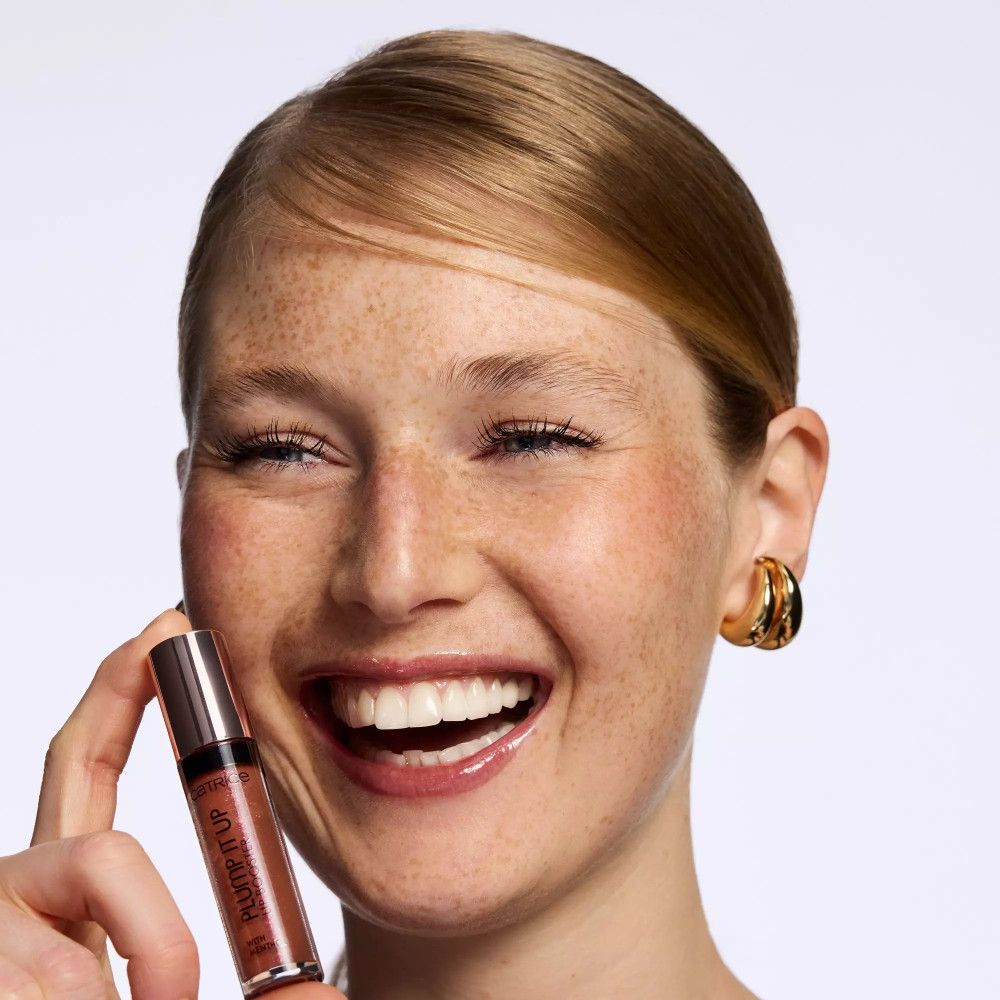 Vrouw houdt Lip Booster vast. Vrouw lacht. Product in hand. Glanzende lippen. Gouden oorbellen.