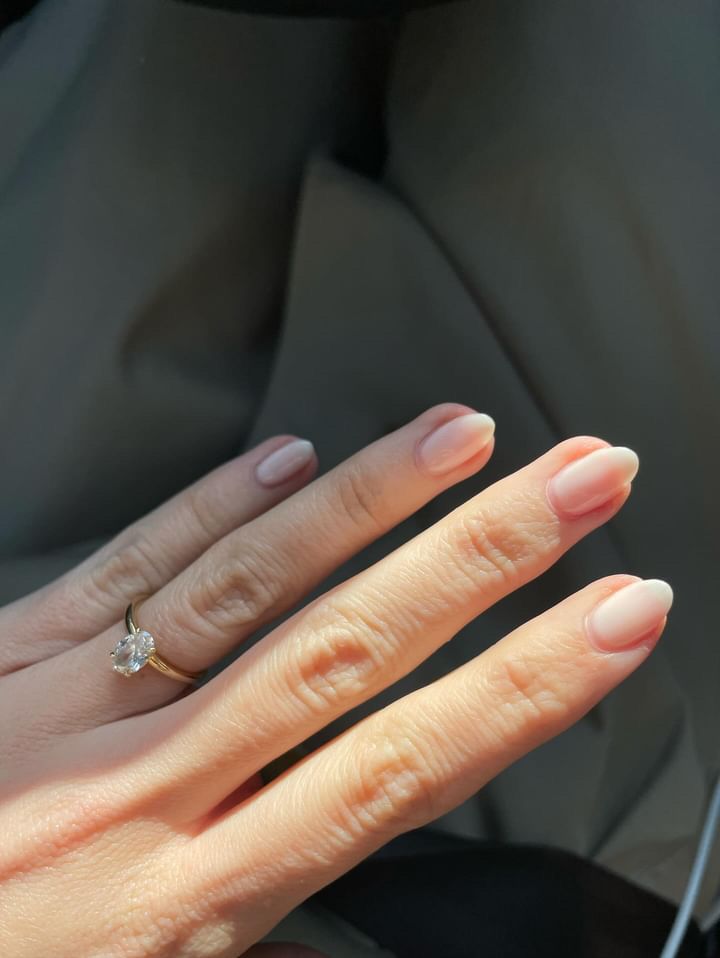 Gros plan d'une main avec des ongles ovales, légèrement vernis. Une bague en or avec diamant au doigt.