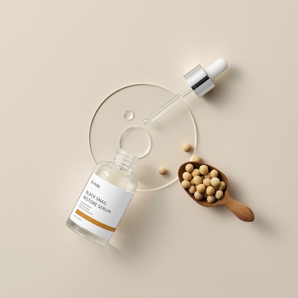 Flacon ouvert avec pipette, à côté d'une cuillère en bois avec des haricots et un disque transparent. Produit : iUNIK Black Snail Restore Serum.