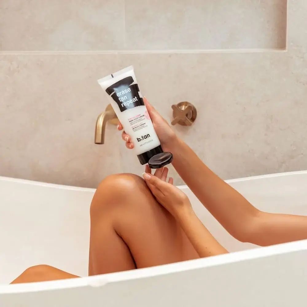 Personne tenant un tube au-dessus d'une jambe dans une baignoire. Le tube porte "erase tan repeat" et "b.tan".